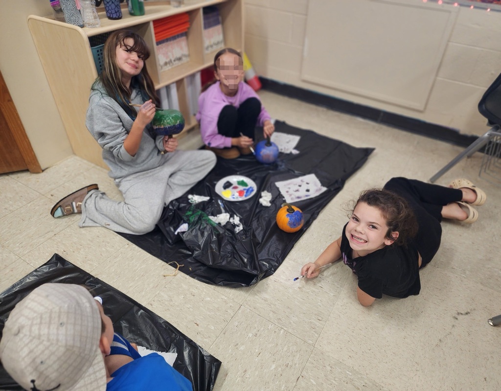 Photos show elementary students participating in a PBIS reward activity by painting pumpkins in various rooms throughout the school. Students use paint and art supplies to decorate pumpkins in different colors and designs while working at tables and classroom spaces. The pumpkins were provided by Moore’s Piggly Wiggly for students who earned enough PBIS points to participate in the fall activity.