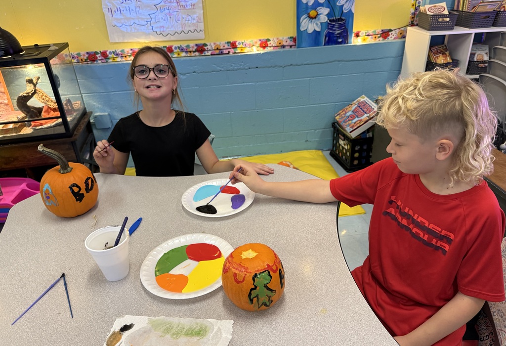 Photos show elementary students participating in a PBIS reward activity by painting pumpkins in various rooms throughout the school. Students use paint and art supplies to decorate pumpkins in different colors and designs while working at tables and classroom spaces. The pumpkins were provided by Moore’s Piggly Wiggly for students who earned enough PBIS points to participate in the fall activity.