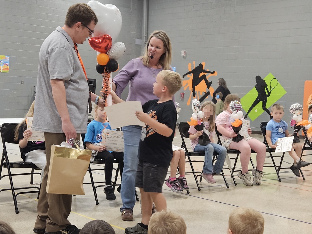 During the Kindness Assembly, a student who earned the most hearts on the school's Kindness Wall receives a special recognition gift. The student stands in front of seated award recipients while accepting a handmade tiger and certificate. Other students holding Kindness Award certificates and balloon bouquets are visible in the school gym as the assembly celebration continues.