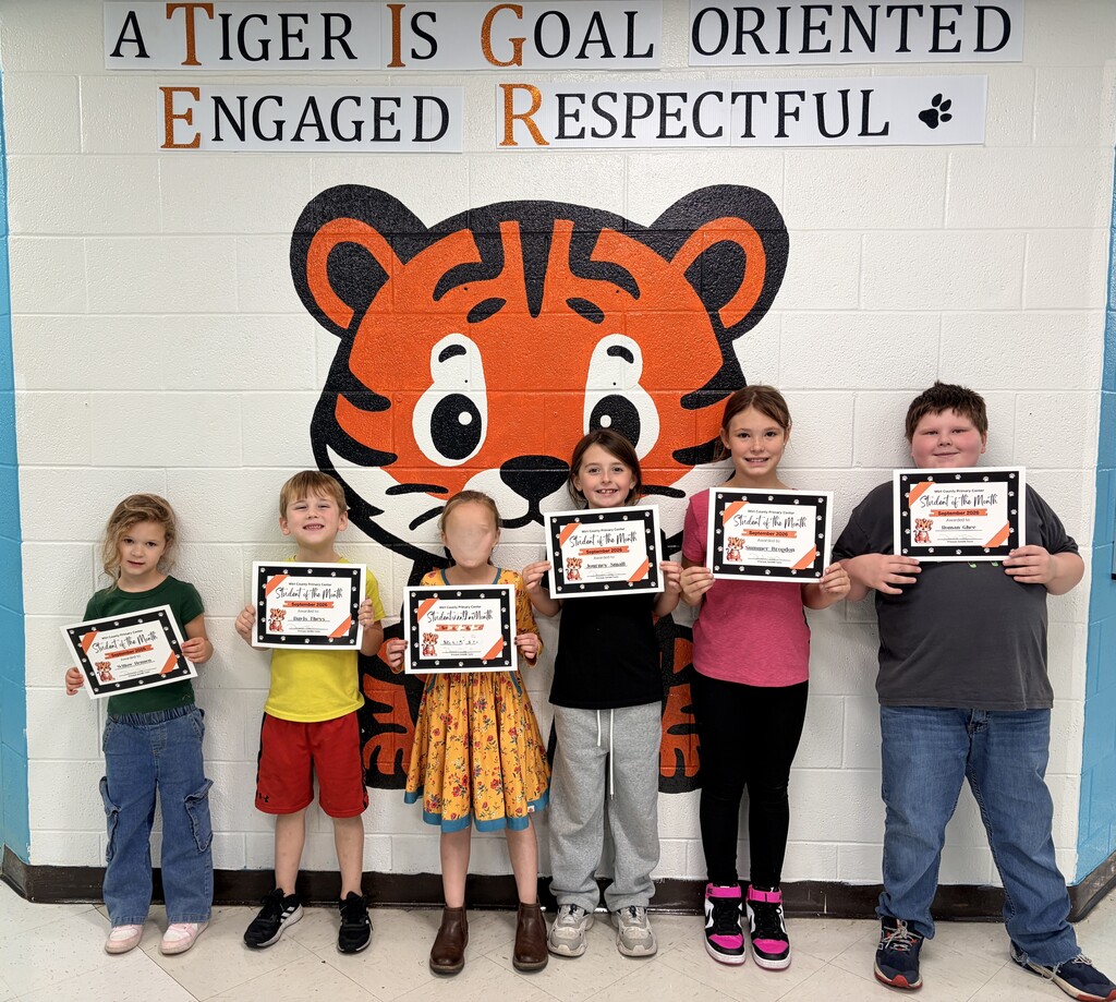 Six Student of the Month recipients from different grade levels stand in front of a large tiger mural at Wirt County Primary Center. Each student holds a Student of the Month certificate. Above the mural are signs displaying the school's character traits, including being goal-oriented, engaged, and respectful. The students are lined up side-by-side in a school hallway.