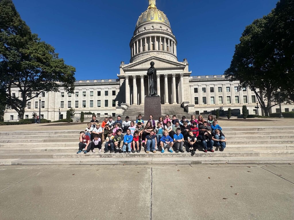 Wirt County Primary Center third grade students sit together on the front steps of the West Virginia State Capitol in Charleston. The Capitol Building, with its large gold dome, white columns, and central statue, stands behind the students under a clear blue sky. Trees frame both sides of the building as the students pose for a group photo during their field trip.