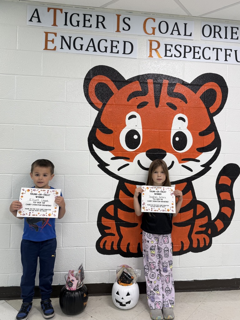 Two students stand in front of a large orange-and-black tiger mural on a school hallway wall. Each student is holding a certificate recognizing them as a Trunk-or-Treat Candy Donation Drawing winner. Two pumpkin-shaped containers filled with prizes are placed on the floor in front of the students. Above the mural are school character words that include “Goal Oriented,” “Engaged,” and “Respectful.”