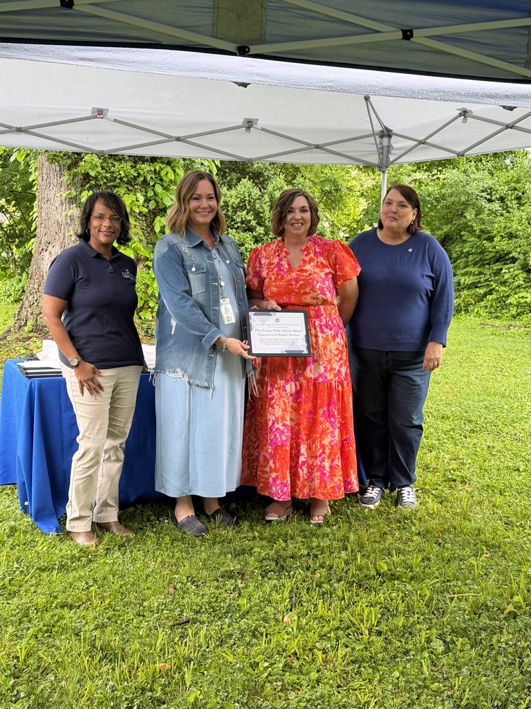 Four people stand under a tent, one holding a certificate. Two tables are behind them. Green grass, trees, and trees in the distance.