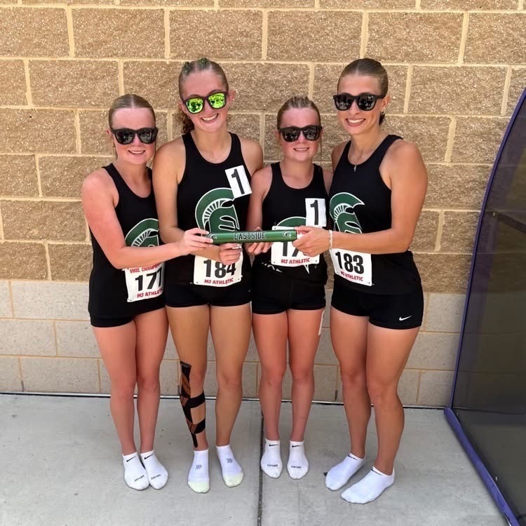 Four women in sports gear hold a baton. They stand in a row, smiling. Behind them is a brick wall.