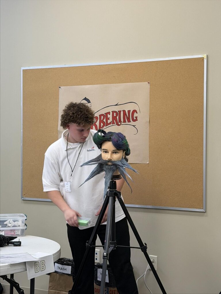 Person with a curly hairstyle working on a mannequin head with a beard and green headpiece.