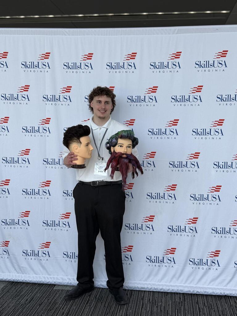 A man in a white shirt and black pants stands with mannequin heads and a doll, against a backdrop with 'SkillUSA' branding.