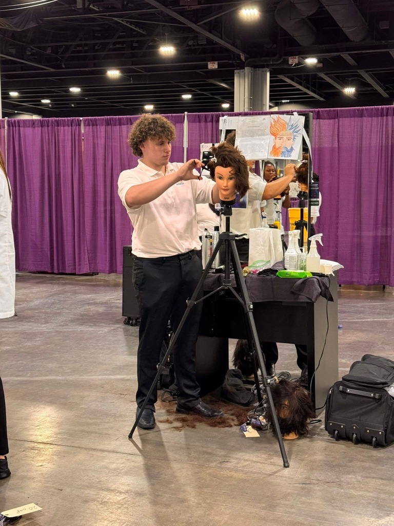 A man behind a tripod camera, taking a photo of a mannequin with curly hair in an indoor area.