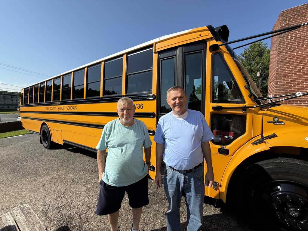 Two men stand in front of a yellow school bus parked in front of a brick building.