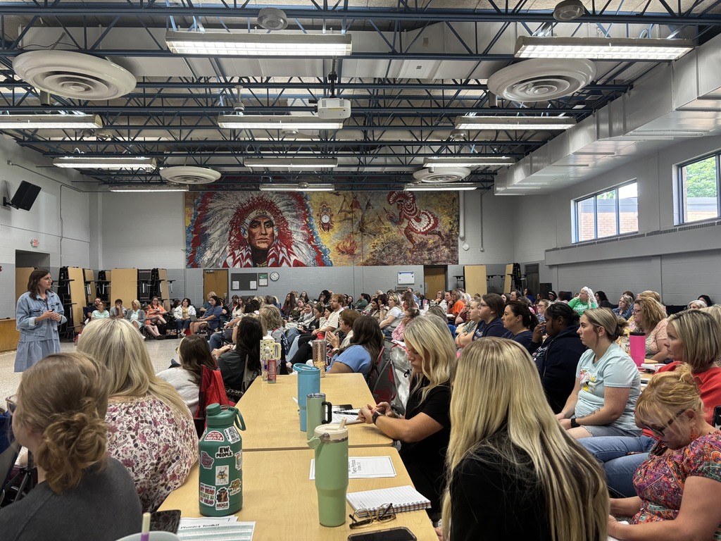 Indoors, people sit around long tables. A woman at a podium speaks. A large painting decorates the wall.