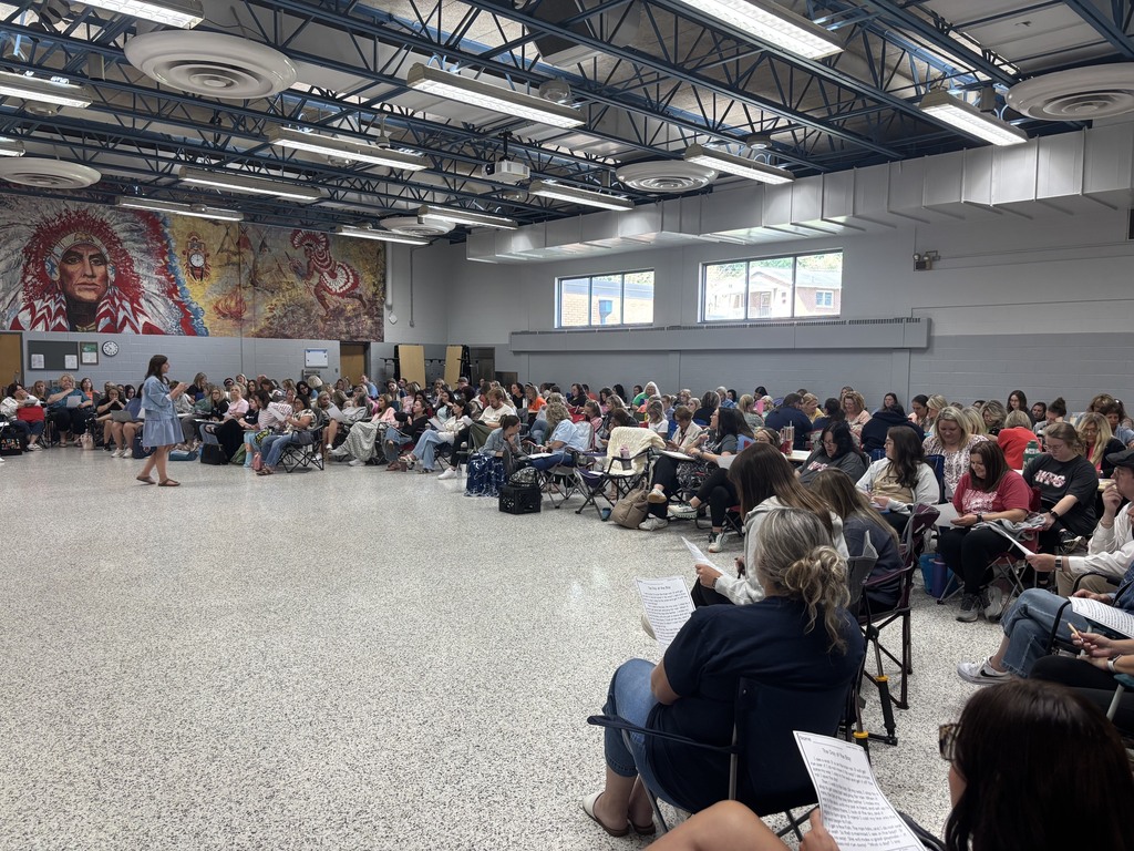 A large room with many people seated and standing. A woman in white walks across the floor.