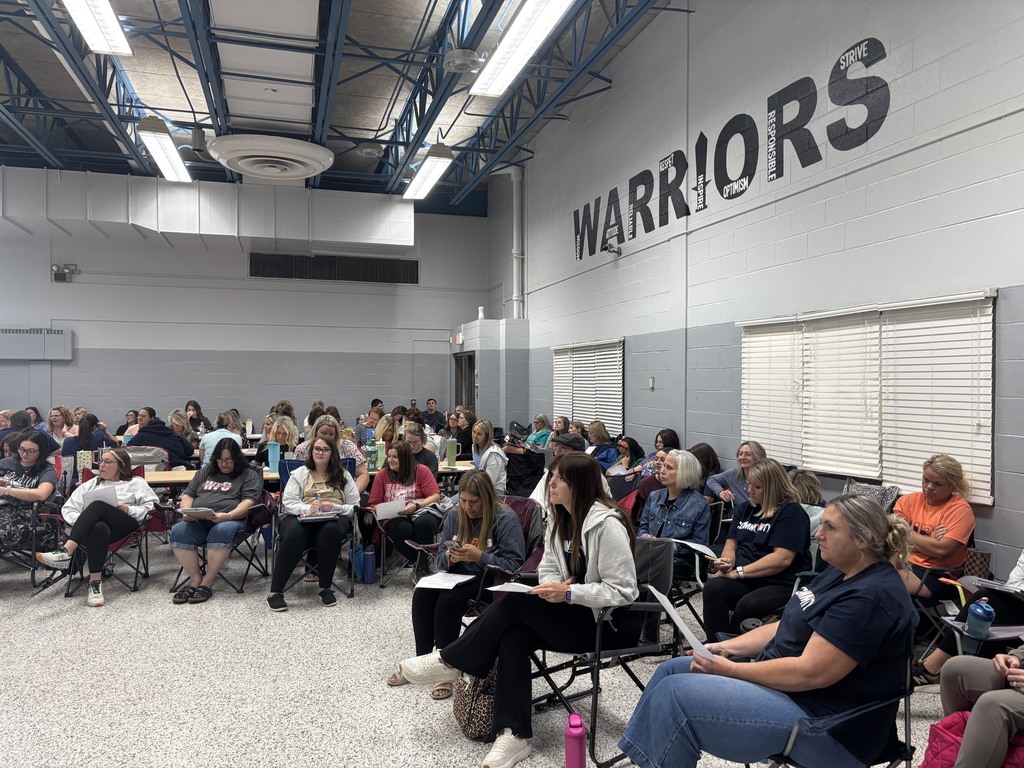 A large group of people sit in a room with white walls, some writing, some listening. A large word, Warriors, is painted on the wall.