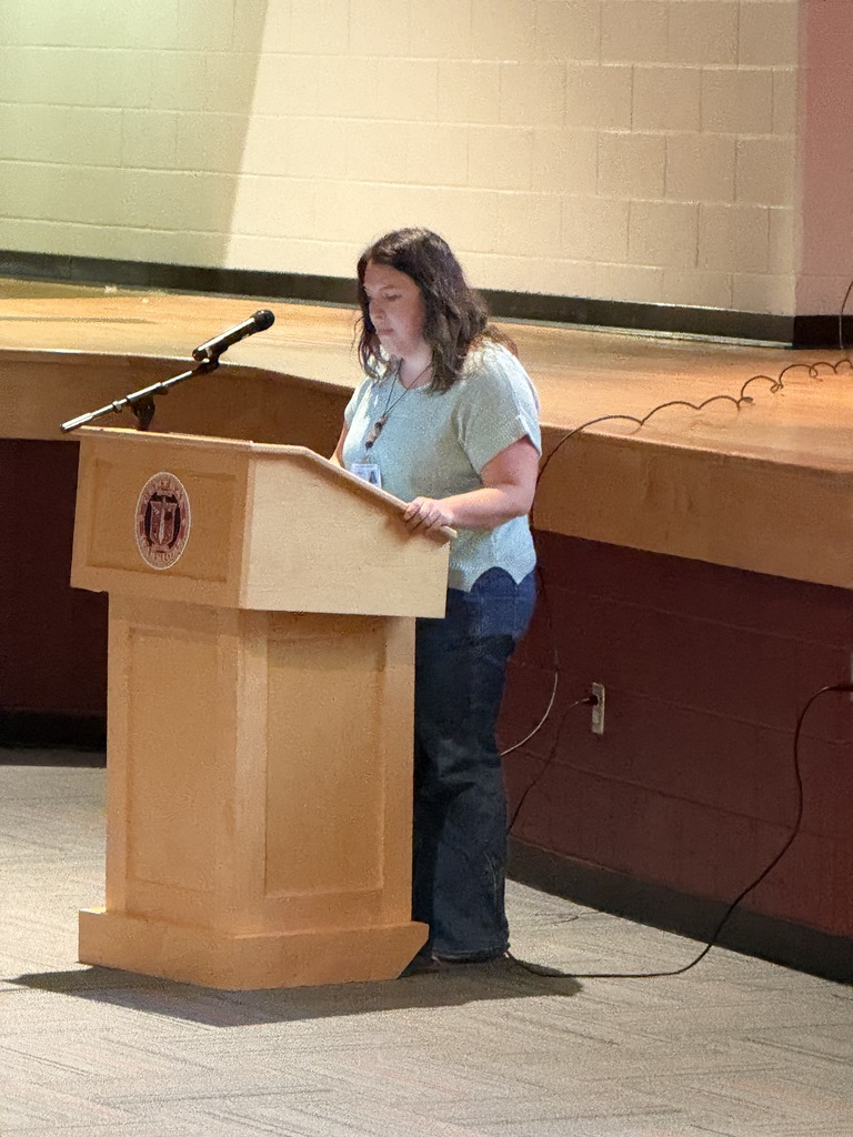 A person stands at a wooden podium with a microphone. A brick wall is behind them.