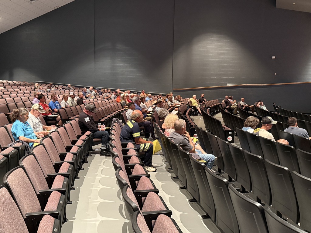 An auditorium with rows of chairs. The chairs are dark blue and maroon. Some people are seated, others stand, and two people wear safety vests.