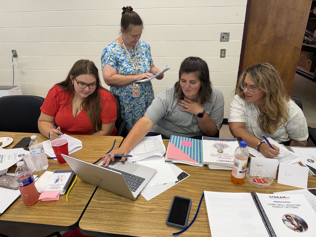 Four women sit around a table, looking at a laptop. One woman wears glasses and holds a pen.