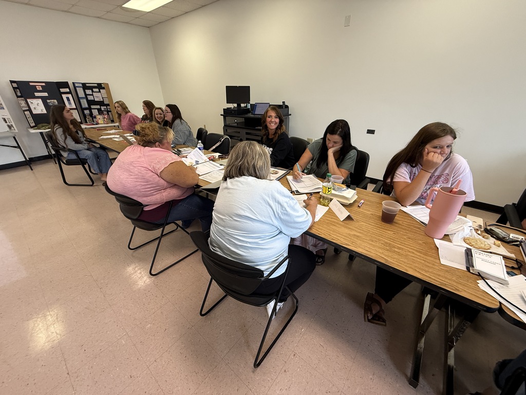 A group of people sit at tables in a room, facing each other. Some have papers and cups.