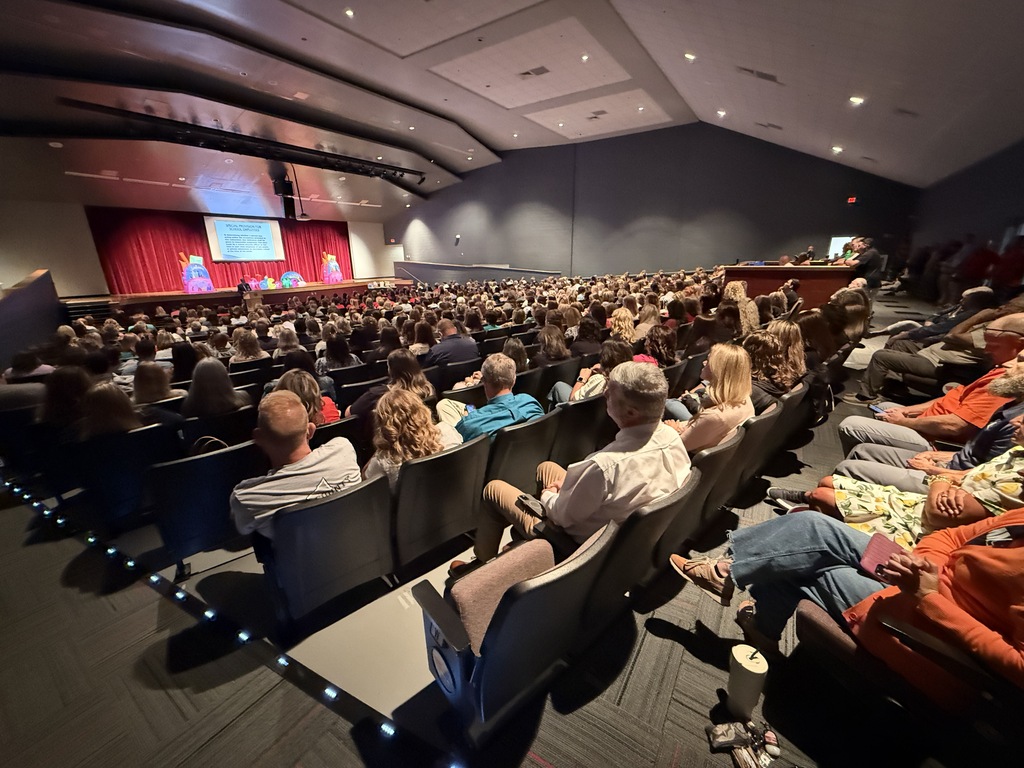 Auditorium with a stage and screen. Many people seated, facing the stage. Red curtain.