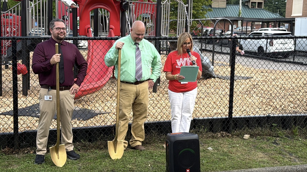Three people stand in front of a fence at a playground. A man holds a shovel. A woman holds a clipboard.