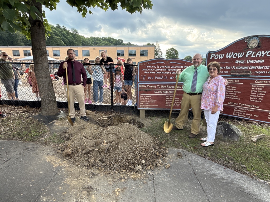 A group of people, including two adults holding shovels, are standing next to a tree and a pile of dirt.