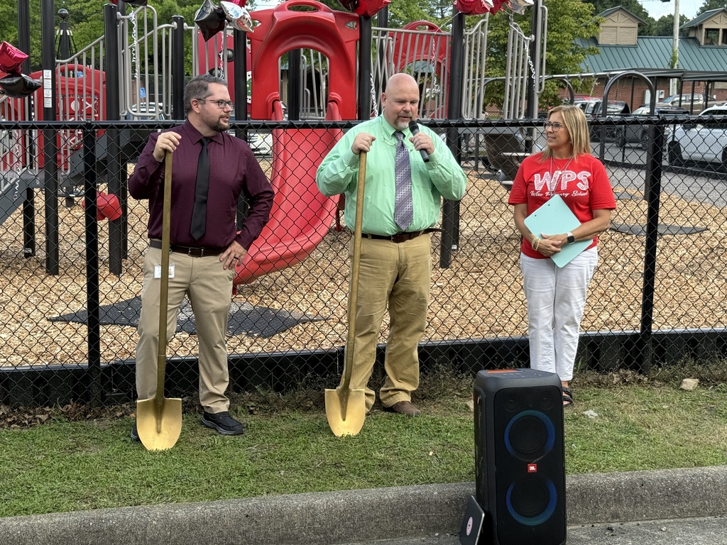 Three people stand in front of a fence, holding shovels, one with a microphone. A playground is behind them.