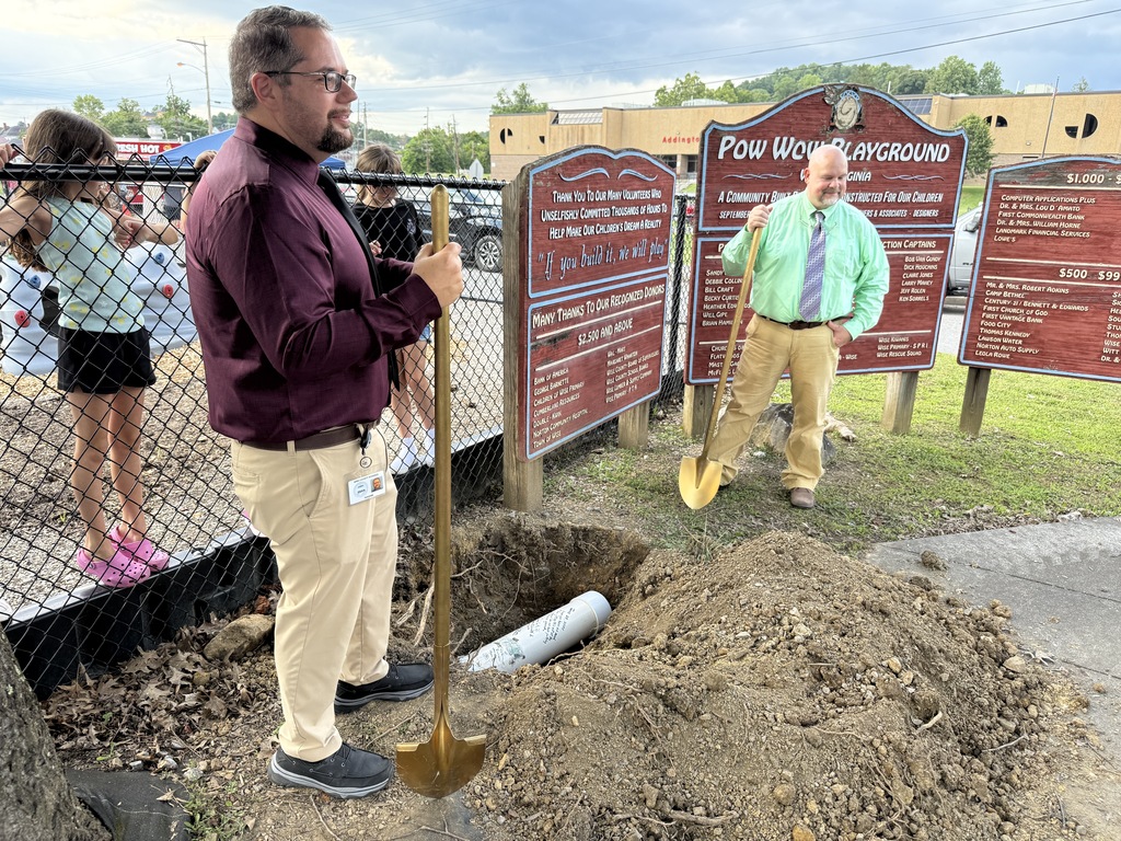 Two men stand on a dirt mound with shovels. Behind them is a fence and a sign.