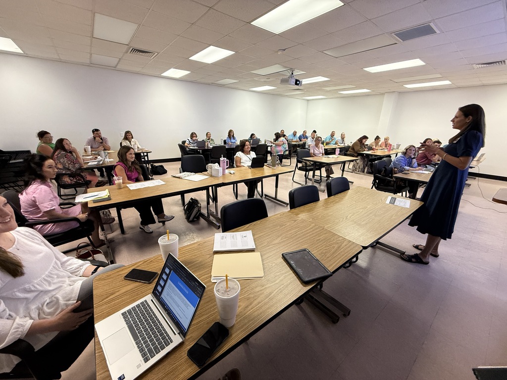 An instructor leads a seated audience in a conference room with tables, laptops, and a projector.