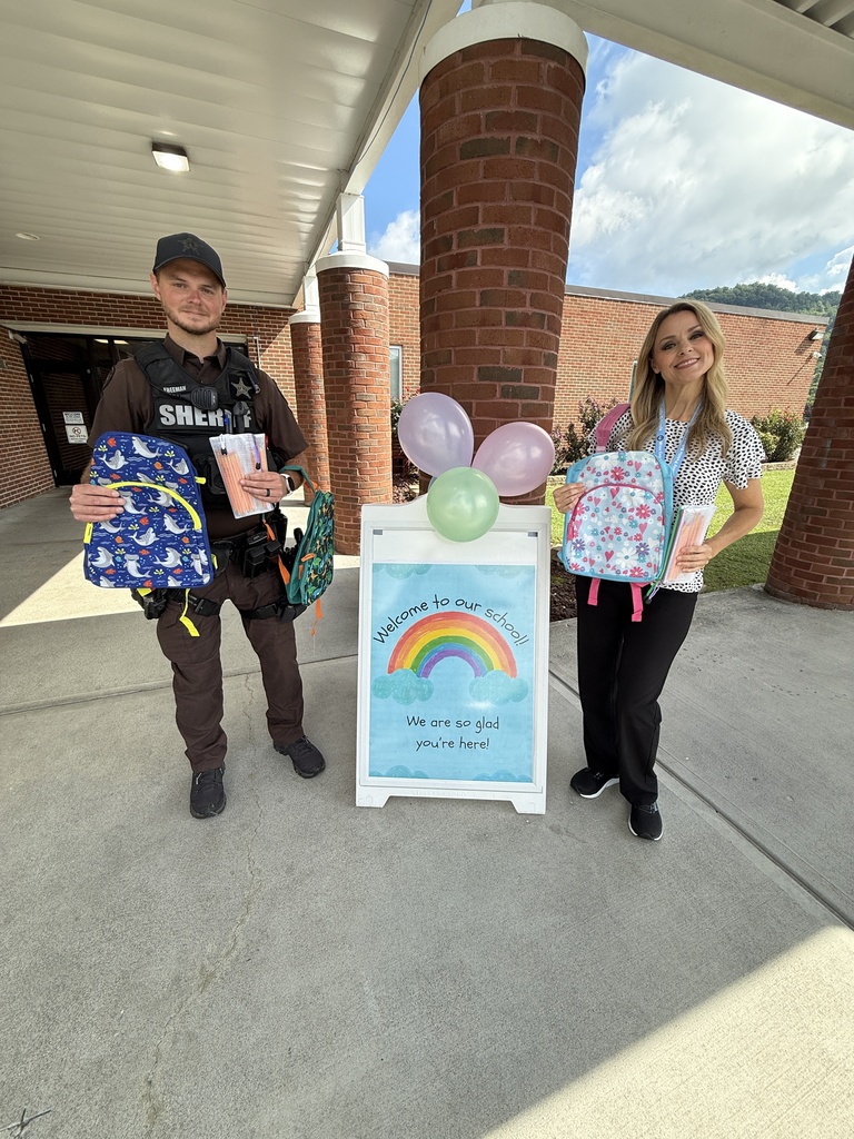 Two people stand outside a building. One holds a backpack, the other holds balloons and a sign with a rainbow.