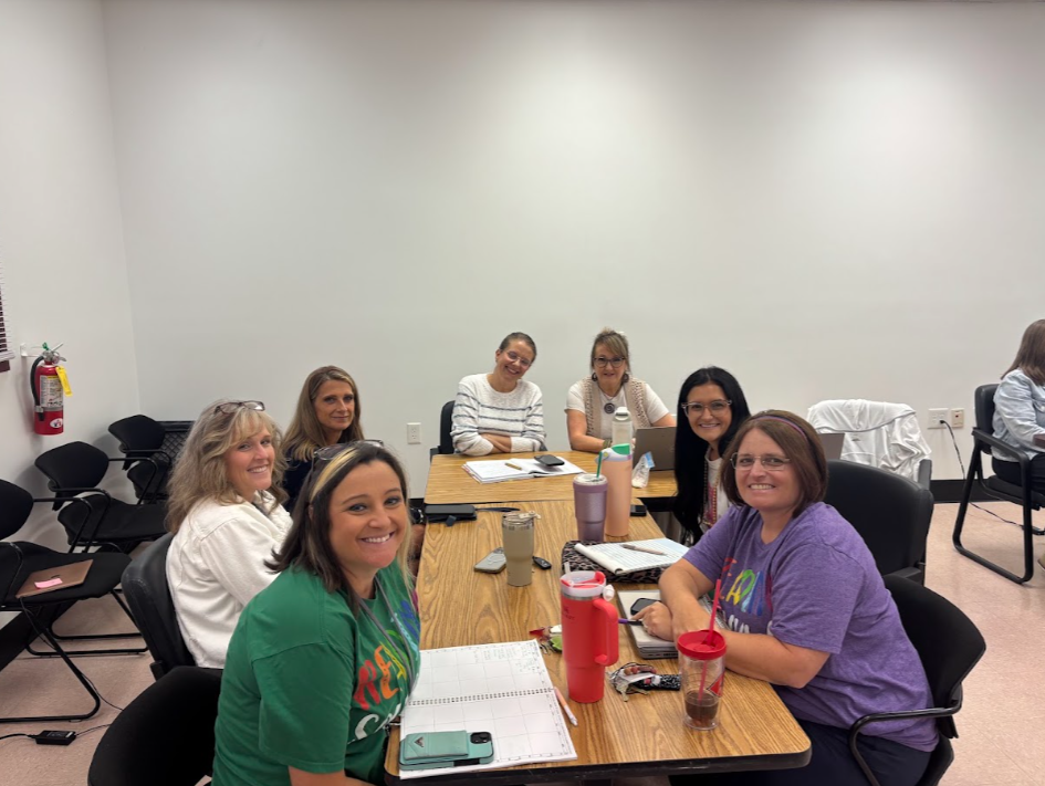 Group of smiling women in a conference room. One has a green shirt, another a purple shirt. Papers, pens, and cups on the table. White walls.