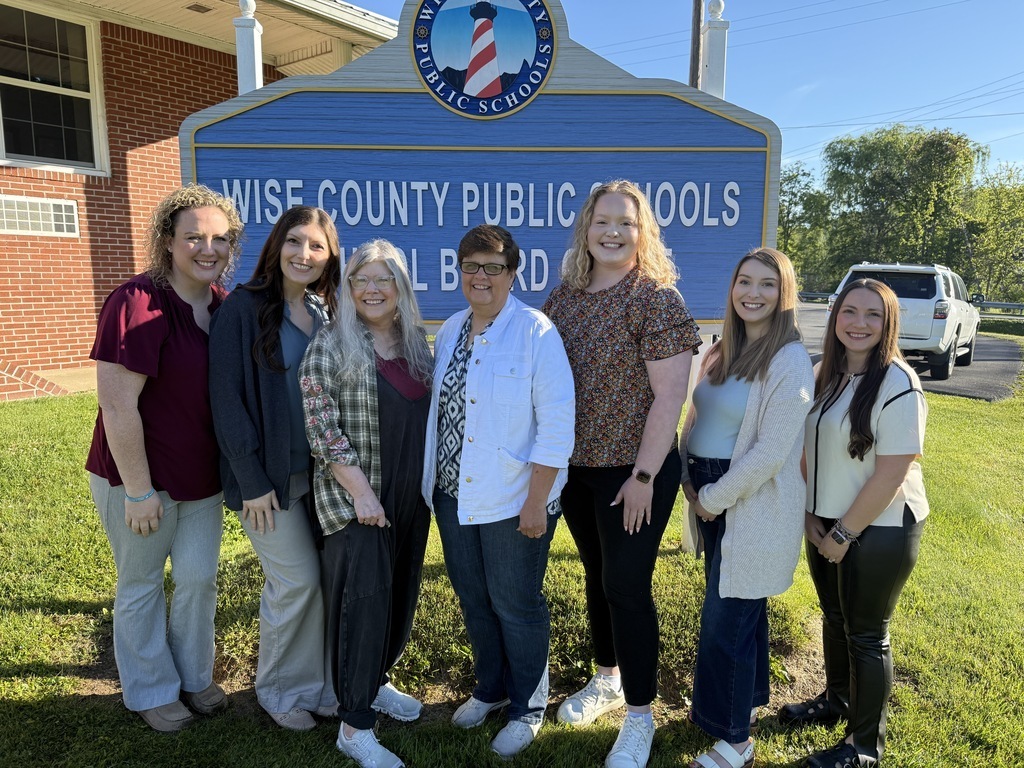 A group of women in business attire standing in front of a blue sign. Background includes a brick building.