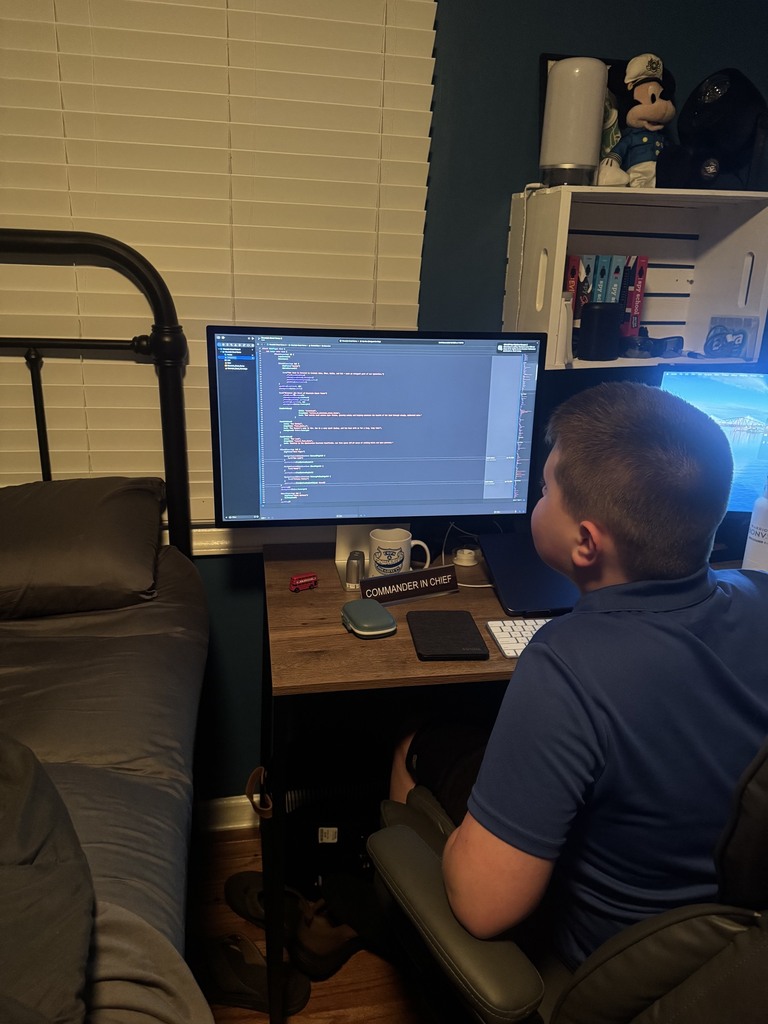 A person sits at a desk with two computer monitors, working on a computer. A bed with a pillow is in the background.