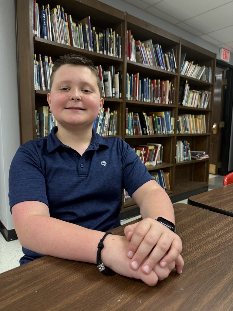 A boy in a navy blue shirt sits at a desk, smiling with bookshelves in the background.