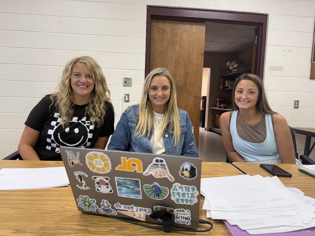 Three women sit around a table with a laptop displaying various stickers, papers, and a phone.