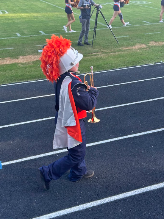 A child in a red feather headdress plays a trumpet on a track, with two cheerleaders and a photographer in the background.