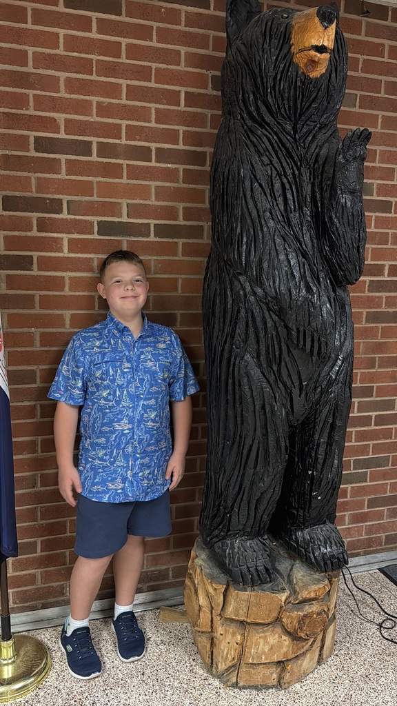 Boy stands next to a large black bear sculpture against a brick wall. The bear is carved from wood.