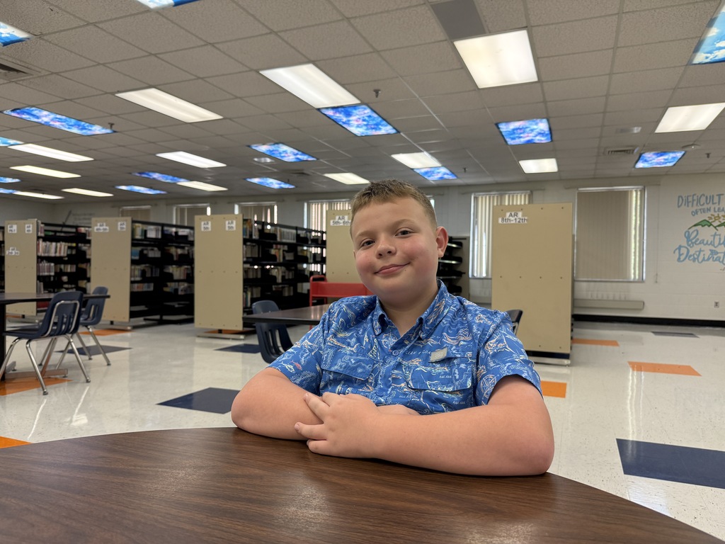 A young boy in a blue shirt sits at a round table in a library with bookshelves and chairs.