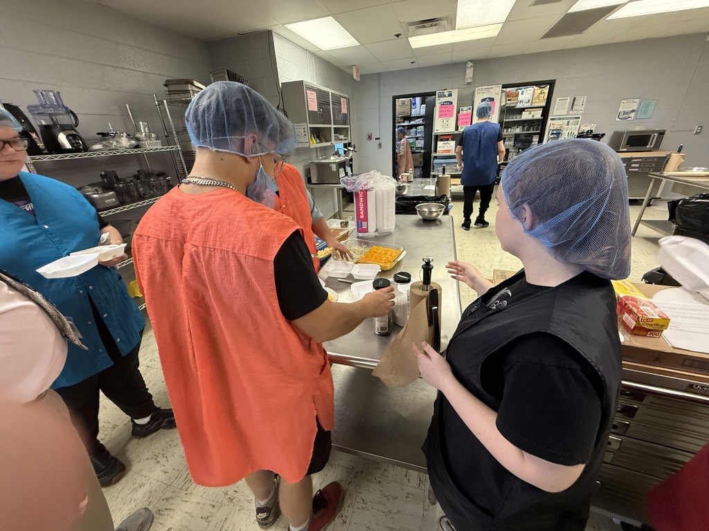 A group of people in a kitchen, wearing hairnets and aprons, prepare food with trays and containers.