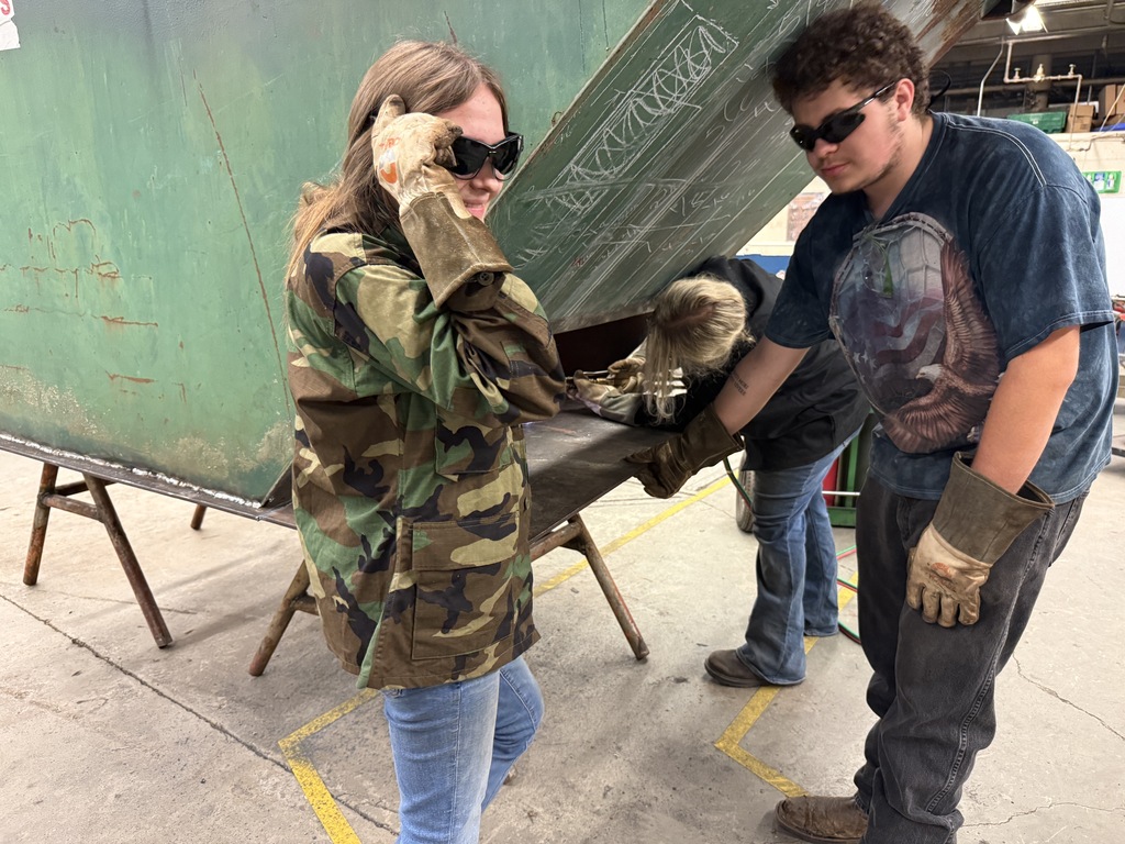 Two people in a workshop with a large green container. One person is wearing gloves and appears to be handling something.
