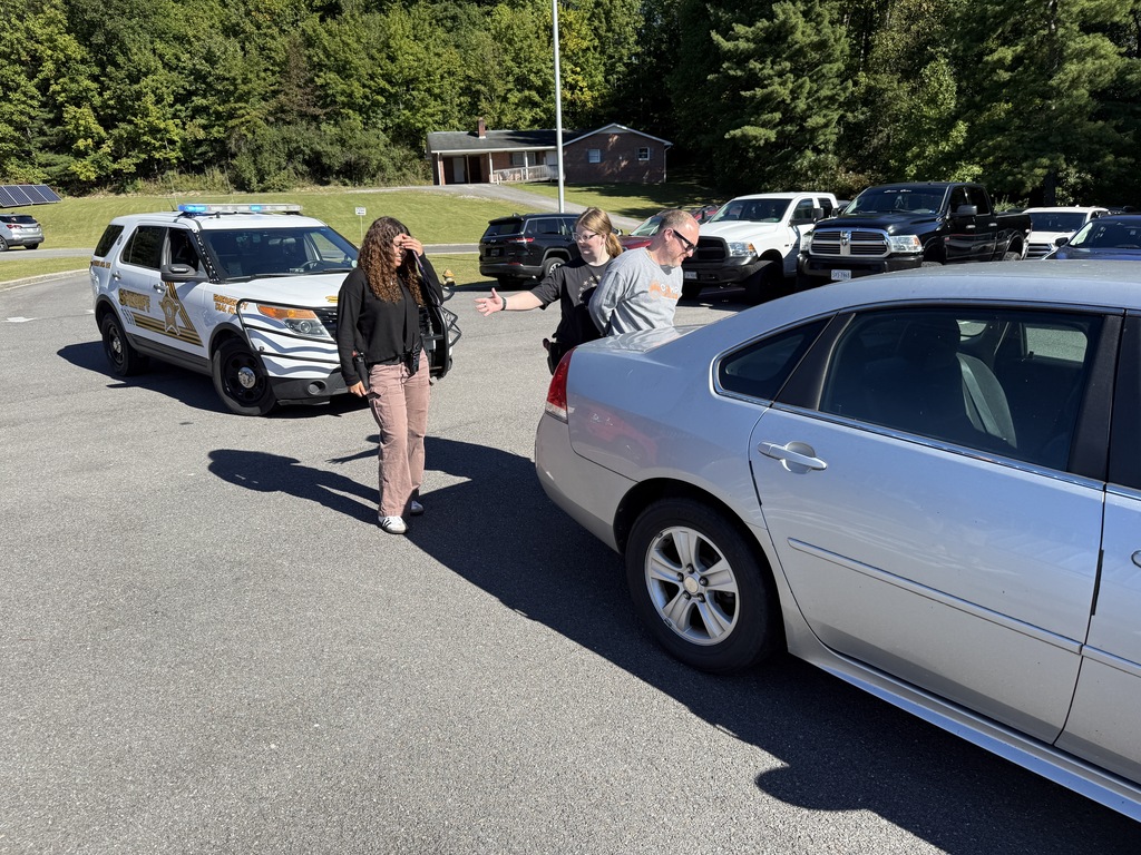 A person assists a woman walking in a parking lot with multiple vehicles parked, including a police SUV.