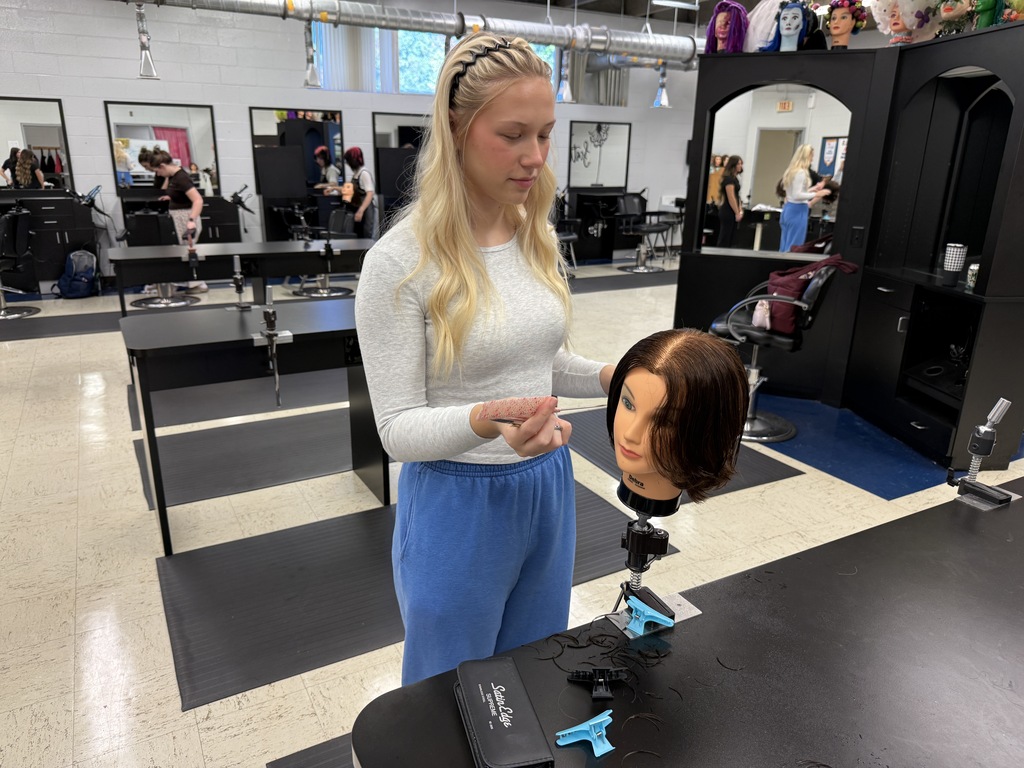 A young woman stands in a salon, adjusting a mannequin's hair while other trainees work in the background.