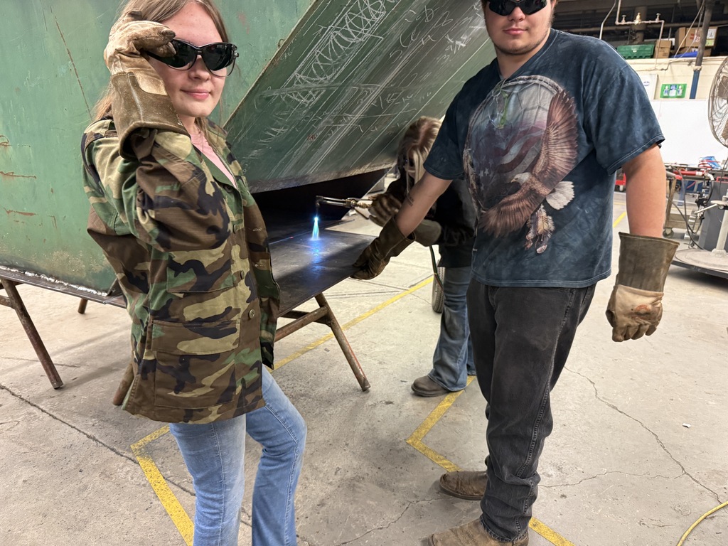 A man and a woman in protective gear are standing by a metal object in a workshop.