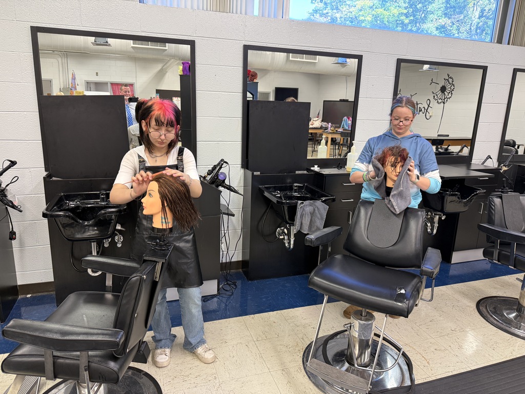 Two people in a salon. One person is standing, holding a mannequin head, and the other is seated in a chair.