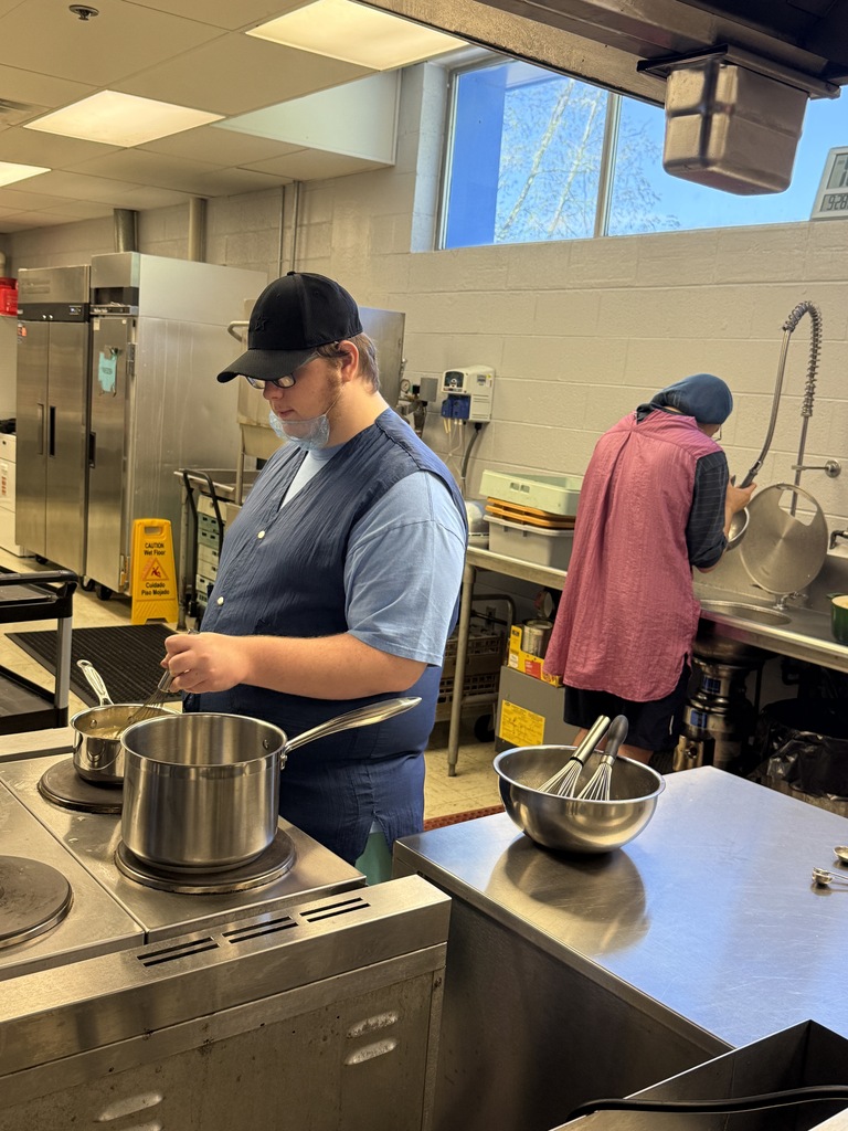 Two people in a commercial kitchen. One prepares food at a stove. Another washes dishes at a sink.