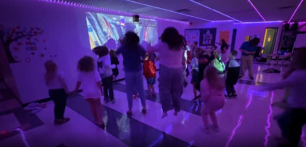 Children dance in a room with purple lighting, a projector screen, and colorful decorations.