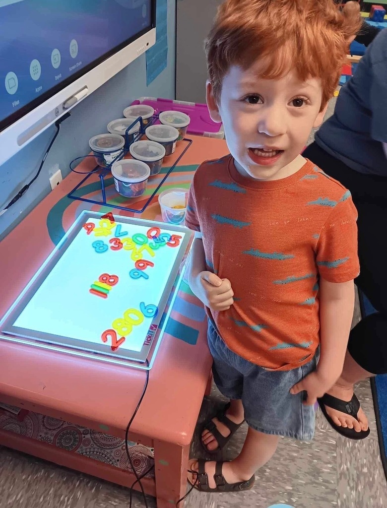 A young child, smiling, stands by a pink table. The table has a lighted screen displaying numbers.