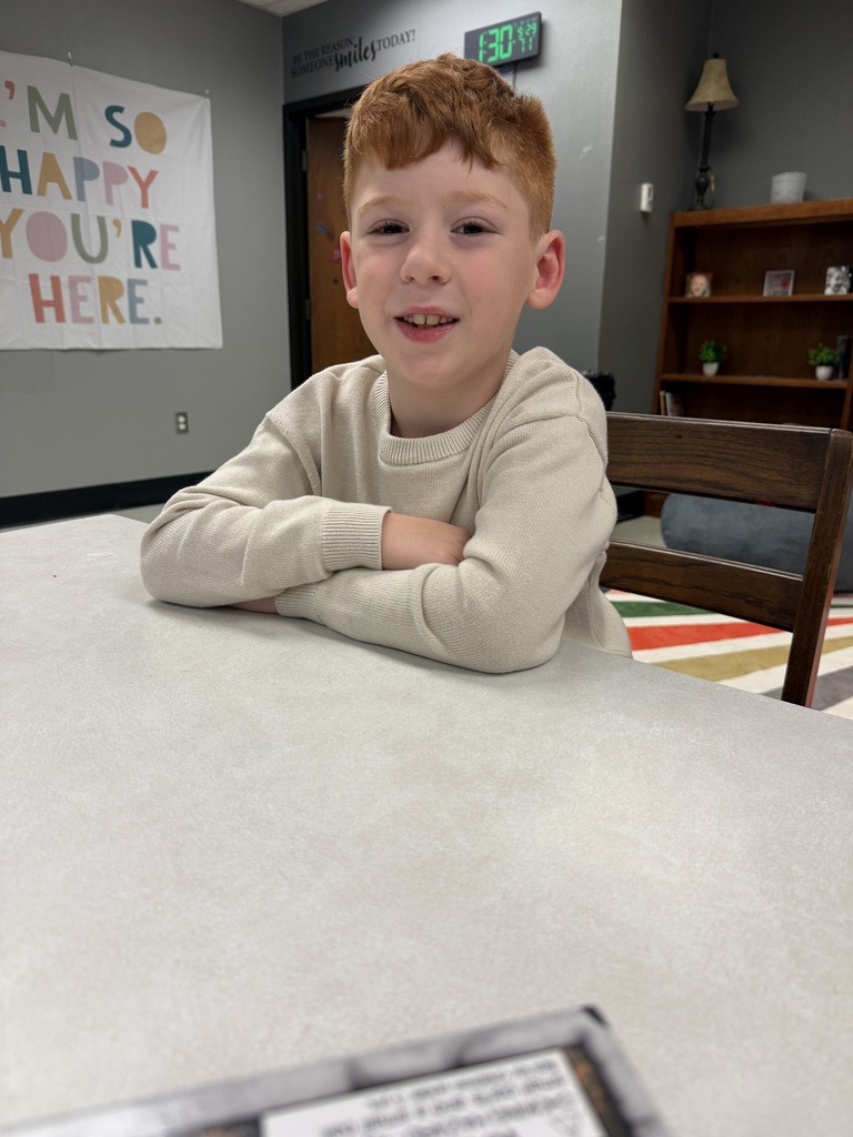 A young boy with red hair sits at a table, smiling with arms crossed, near a banner and bookshelf.