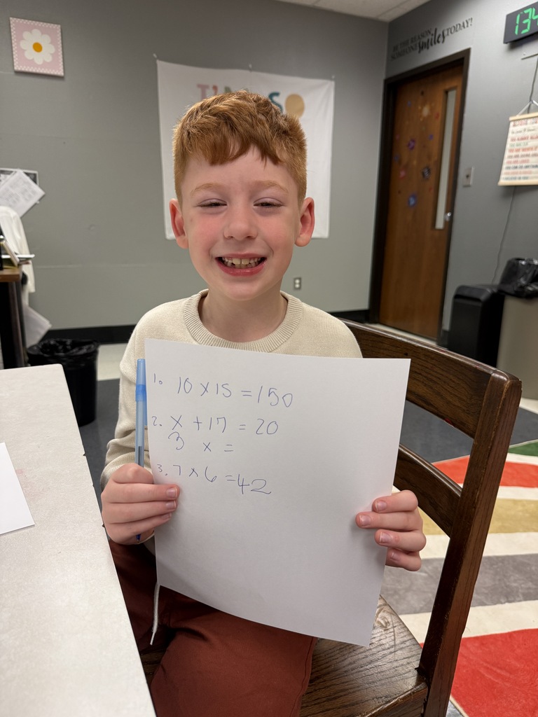 A child sits in a chair, smiling, holding a paper with written mathematical equations. Behind, a door, papers on the wall, and a trash can are visible.
