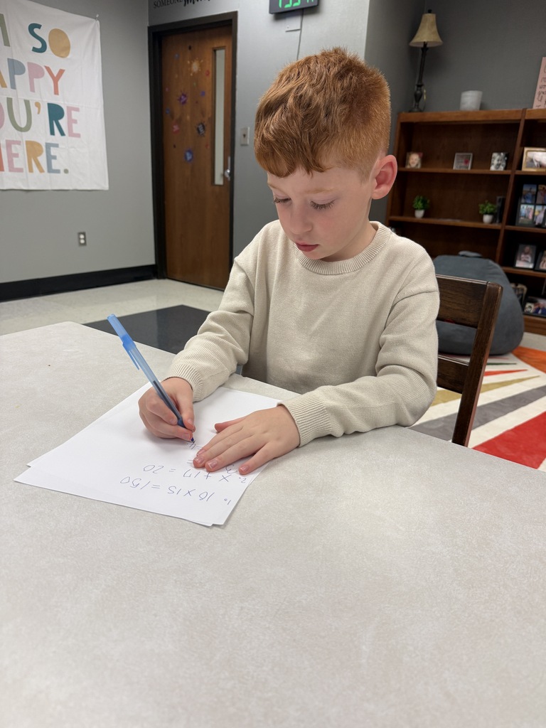 A young boy writes on a piece of paper with a pen at a table. A banner on the wall reads "So happy you're here".
