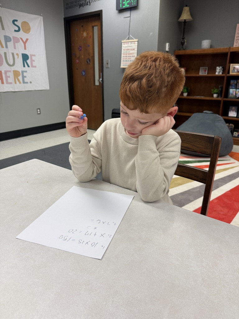A child is seated at a table, focusing on a piece of paper. A sign on the wall reads "So Happy You're Here."