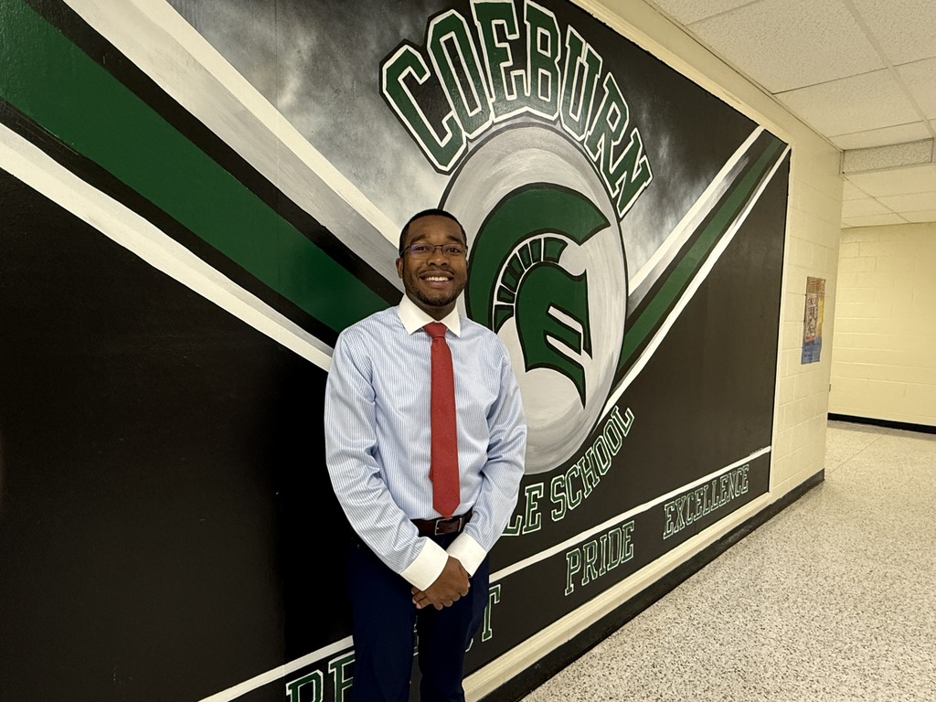 A man in a red tie stands in front of a wall with a large school logo.