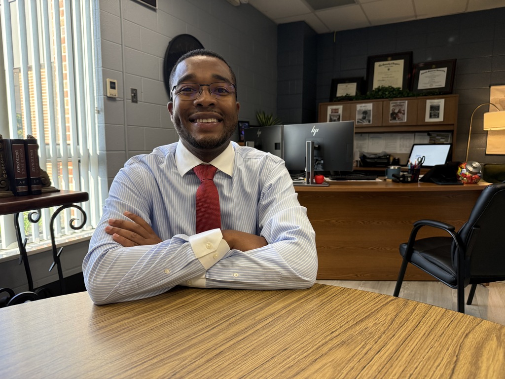 A smiling man in a light blue shirt and red tie sits in an office, arms crossed.