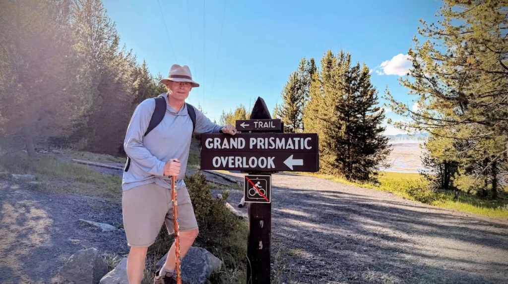 Person holding cane by a sign reading Grand Prismatic Overlook, surrounded by trees, and a body of water.